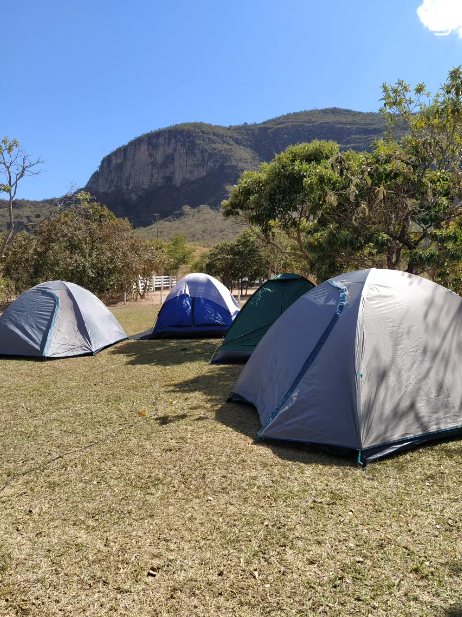 Barracas espalhadas no gramado do camping sob céu azul, com o paredão ao fundo